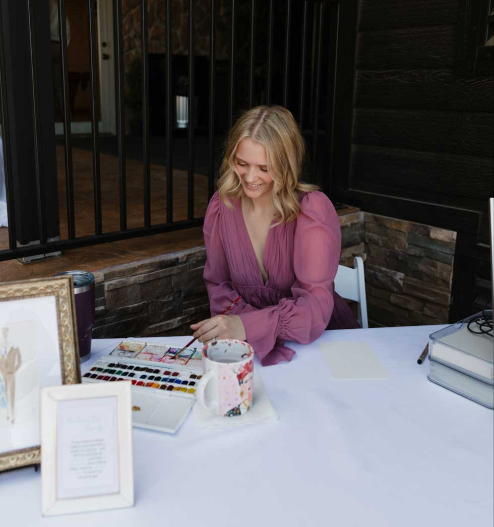 Woman in a pink dress sitting at a table with art supplies in a rustic setting